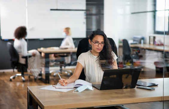 Hispanic Program Evaluator Wearing Eyeglasses sitting at her desk in an Office working on a laptop 
