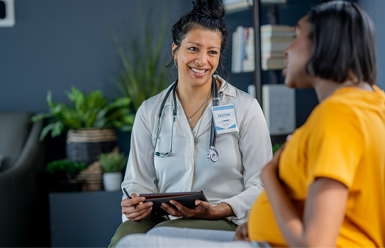 Midwife wearing lab coat and stethoscope holding paper and pen looks caringly at pregnant woman wearing a yellow dress and talking about her pregnancy
