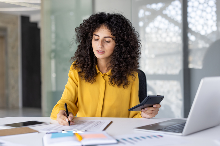 A focused professional woman is meticulously reviewing financial documents in a modern office setting, holding a calculator and pen.