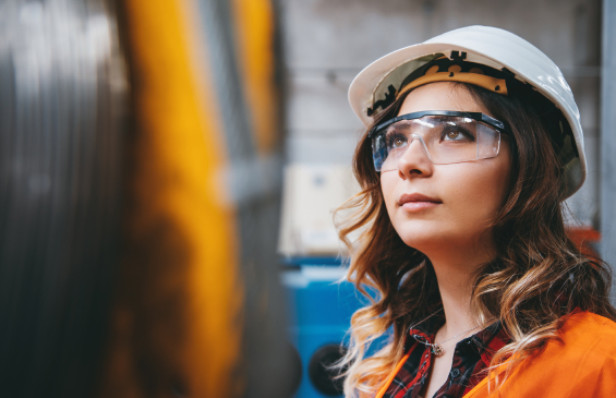 A close-up portrait of an occupational health and safety worker wearing a hard hat and safety glasses looking towards an object.
